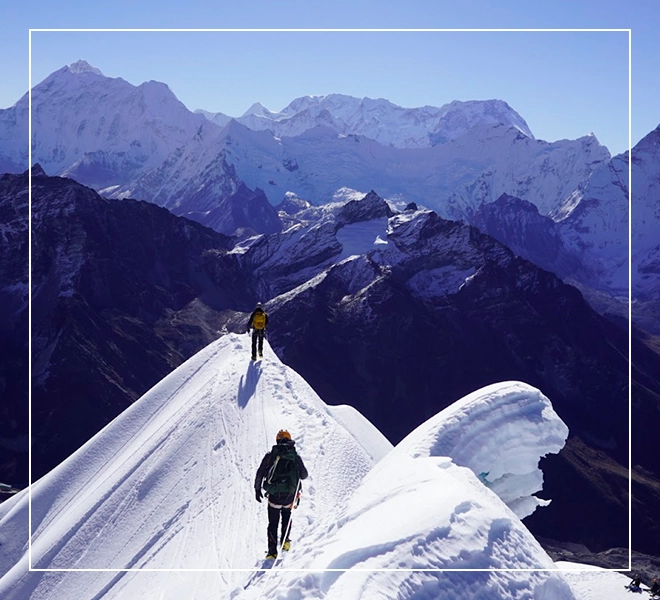 Two people skiing on a mountain top with snow capped mountains in the background.